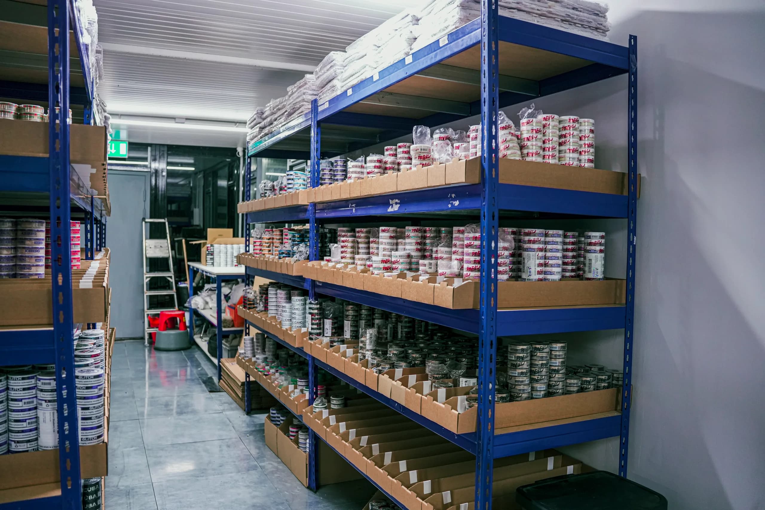 Blue warehouse racking filled with cardboard trays of nicotine pouch tins, stacked several shelves high, with a step ladder further down the aisle